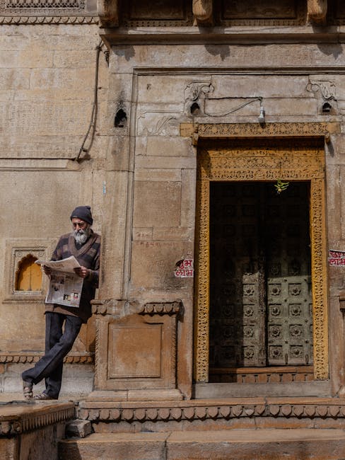 An elderly man reads a newspaper by an ornate doorway in Jodhpur, Rajasthan, capturing architectural beauty.